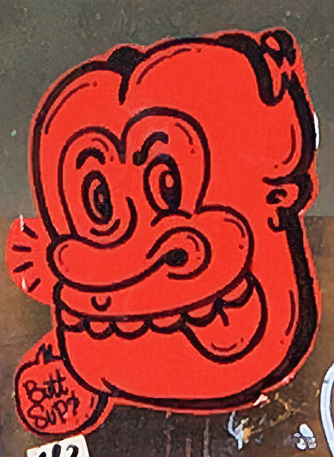 A graffiti sticker of a cartoon face in red and black with the tag "butt sup"