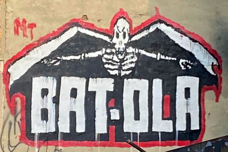 A winged skeleton looms over the words bat-ola in black and white all surrounded with a red border.