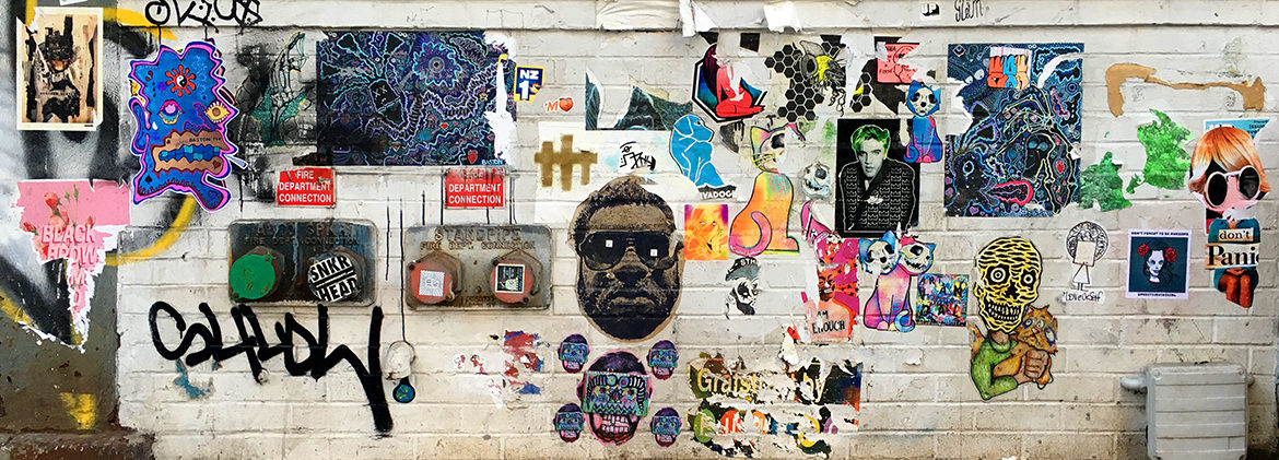 A white wall gallery filled with paste up art illustrations, paintings and stickers